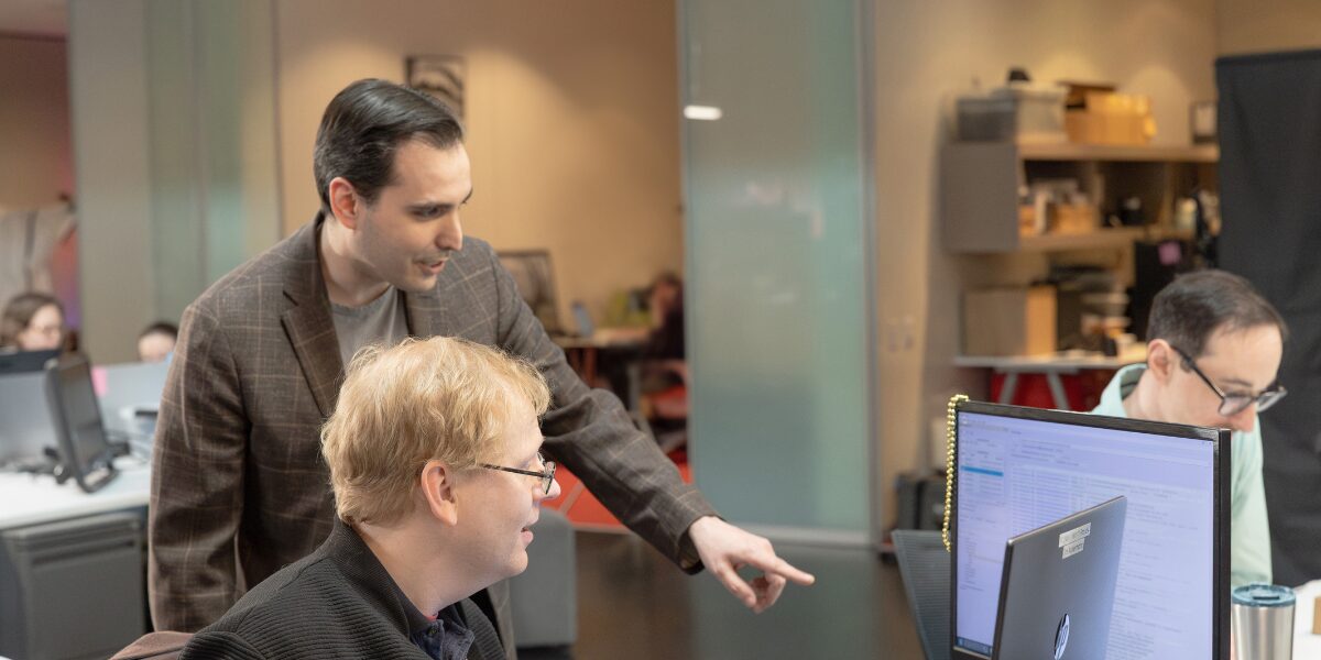 A male Aspiritech team member stands beside a second team member and points at something on a computer screen.