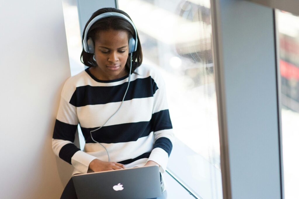 A young woman wearing headphones sits on a windowsill while working at a laptop.