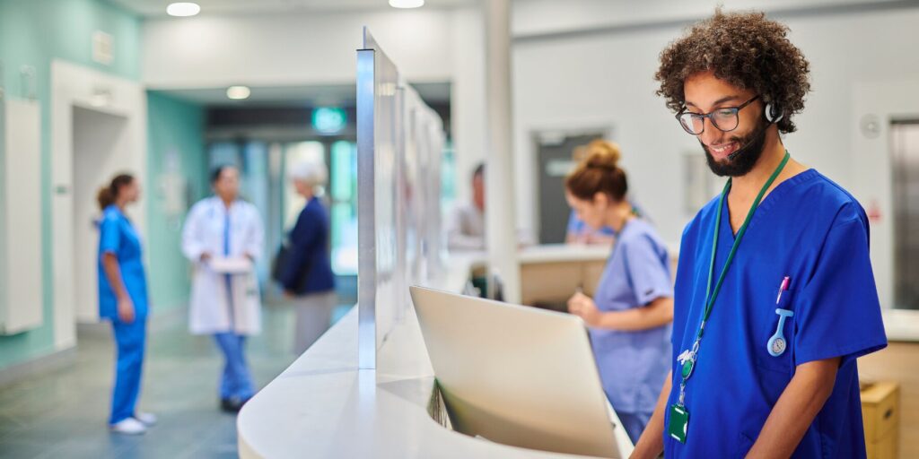 A male medical professional looks at a laptop computer while other medical workers in scrubs and lab coats work in the background.