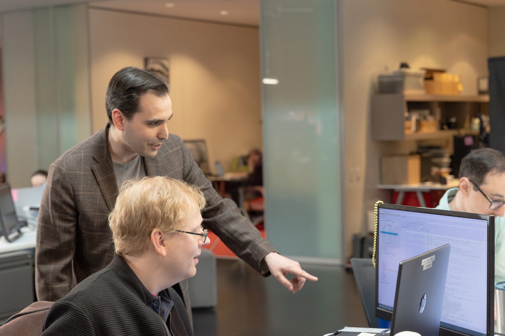 A male Aspiritech team member stands beside a second team member and points at something on a computer screen.
