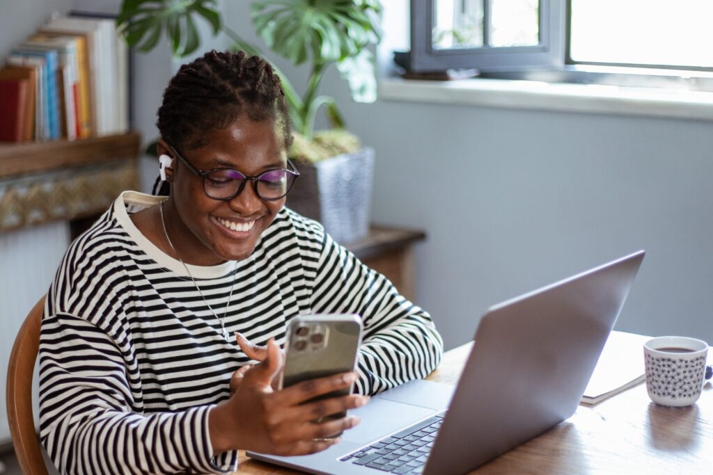 A woman smiles while holding a smartphone and working at a laptop.